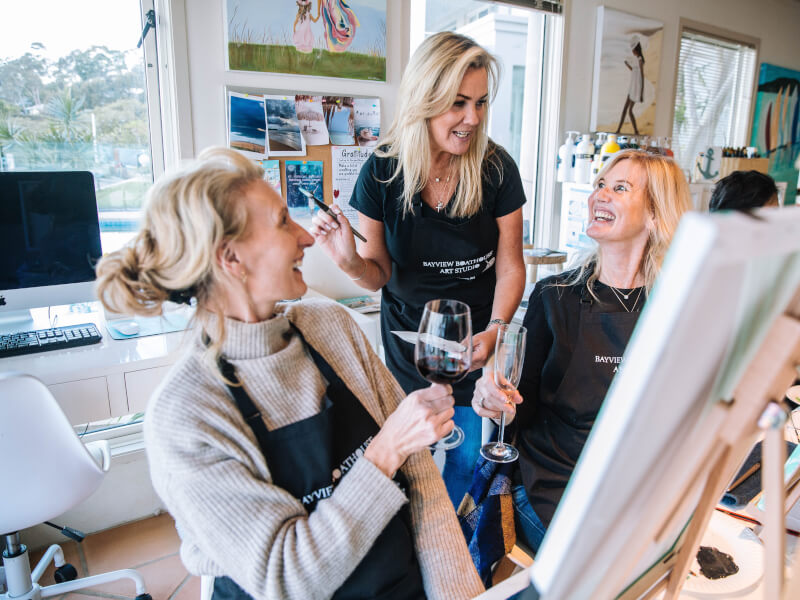 An art teacher smiling at two women at a paint and sip class.